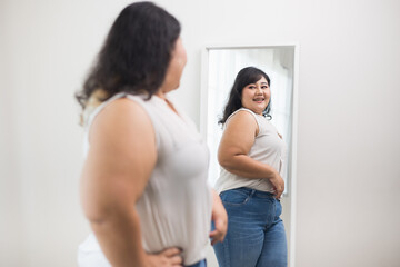 Fototapeta premium Body positive plus size female clothing. Body positive plus size woman happily and is proud of herself looking at mirror at home. Try wearing clothes that measure the size.