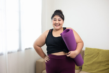 Fototapeta na wymiar Attractive asian young fitness woman plus size holding yoga mat workout at home in living room. Female training and exercise wearing sport wear fit body. Happy and is proud of herself.