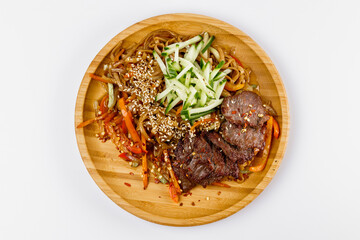 Fried noodles with meat, cucumbers, carrots and sesame seeds with vegetables on a wooden plate. White background.