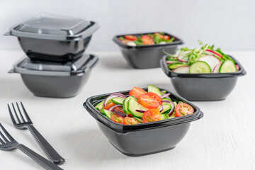 Variety of vegetarian healthy vegetable salads made of sliced cucumber, tomatoes and radish served in black plastic bowl and fork on white wooden background. Food delivery concept and healthy eating