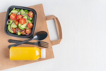 Top view of vegetarian salad made of sliced cucumber and tomato served in black plastic bowl with bottle of orange fruit juice and fork on delivery paper bag on white wooden background with copy space