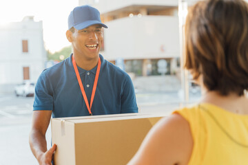Young, smiling Latino delivery man delivering a package at dusk to an unrecognizable woman at her front door.