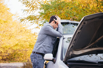 A man looks under the open hood of a car. The car broke down on the road.