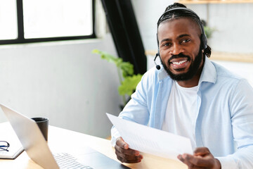 Portrait African American man in headset working remotely at home using laptop. Employee working online analyzing sales report while sitting at home looks at the camera and smiles friendly