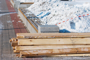 A pile of boards with protruding nails. Winter outdoors. Dismantling of temporary New Year's ice slides after the holiday. A snowdrift of snow on a granite bench with a flower bed. Selective focus.