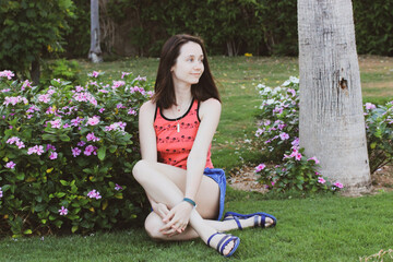 White brunette smiling young woman, student girl in a pink tank top, shorts sitting on green lawn in summer day, enjoying the outdoors. Female in spring garden. Relaxed person being at one with nature - Powered by Adobe