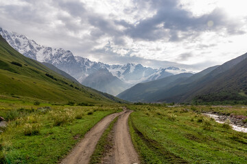 Fototapeta na wymiar A hiking path leading to the Shkhara Glacier in the Greater Caucasus Mountain Range in Georgia, Svaneti Region, Ushguli. Snow-capped mountains in the back. Wanderlust. Wilderness. Overcast and clouds