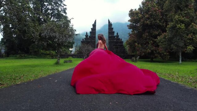 Pretty asian woman runs to balinese gates in long red dress and drone follows in slowmotion on Hangara gates in Bali Indonesia. fashion concept. High quality fullhd aerial footage