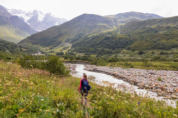 A female hiker walking along the Patara Enguri River flowing down the a valley with view on the Shkhara Glacier in the Greater Caucasus Mountain Range in Georgia,Svaneti Region,Ushguli.Blooming flower