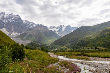 Fototapeta na wymiar Patara Enguri River flowing down the a valley with view on the Shkhara Glacier in the Greater Caucasus Mountain Range in Georgia, Svaneti Region,Ushguli. Snow-capped mountains in the back. Wilderness.