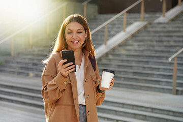 Smiling young business woman holding smartphone and take away coffee in the morning with blurred...