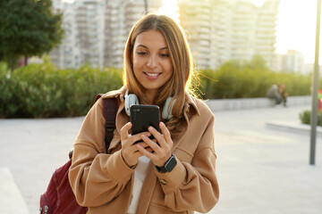 Sunshine girl with coat and backpack chatting with smartphone app on modern city street at sunset. Sustainable living. Millennial generation. Headphones music. Digital native. Smart city.