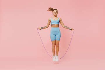 Full body young sporty athletic fitness trainer instructor woman wear blue tracksuit spend time in home gym using skipping rope isolated on pastel plain light pink background. Workout sport concept. - obrazy, fototapety, plakaty