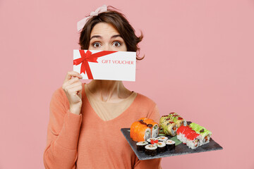 Young woman in sweater hold in hand makizushi sushi roll served on black plate traditional japanese food cover mouth with gift certificate coupon voucher card isolated on plain pastel pink background