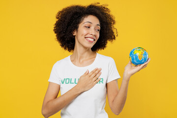 Young woman of African American ethnicity wears white volunteer t-shirt hold in palms Earth world globe put hand on heart isolated on plain yellow background. Voluntary free work help grace concept.