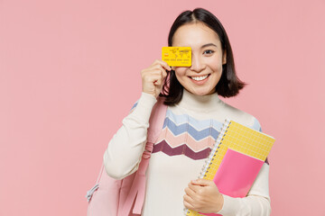 Teen student girl of Asian ethnicity wearing sweater backpack hold books cover eye with credit bank card isolated on pastel plain light pink color background Education in university college concept.