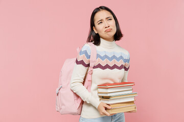 Disappointed sad teen student girl of Asian ethnicity wearing sweater backpack hold pile of books look camera isolated on pastel plain light pink background Education in university college concept
