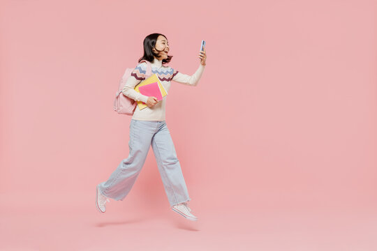 Full Size Teen Student Girl Of Asian Ethnicity In Sweater Hold Backpack Jump High Use Mobile Cell Phone Jump High Isolated On Pastel Plain Light Pink Background Education In University College Concept
