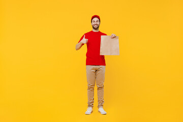 Fototapeta premium Full size delivery guy employee man in red cap T-shirt uniform workwear work as dealer courier hold brown clear blank craft paper takeaway bag mock up show thumb up isolated on plain yellow background