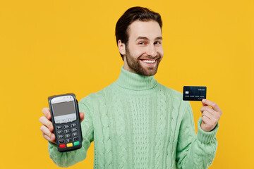 Young man 20s in mint knitted sweater hold wireless modern bank payment terminal to process acquire credit card payments isolated on plain yellow background studio. People lifestyle fashion concept.