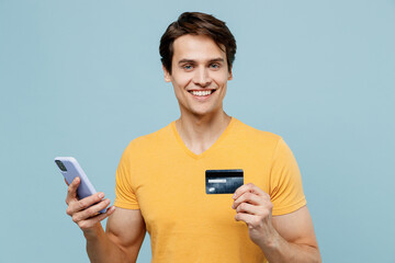Young smiling man 20s in yellow t-shirt using mobile cell phone hold credit bank card doing online shopping order delivery booking tour isolated on plain pastel light blue background studio portrait