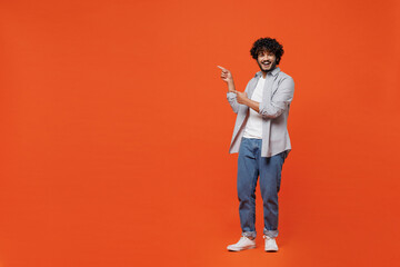 Full size body length fun young bearded Indian man 20s years old wears blue shirt pointing index fingers aside on workspace area copy space mock up isolated on plain orange background studio portrait.