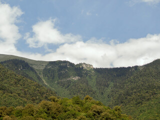 A green mountain range with forested rocks against a blue sky and clouds. Mountain valley on a summer day