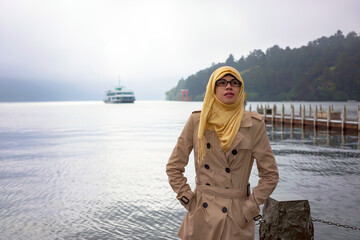 Portrait of muslim woman looking at sky at Lake Ashi in Hakone, Japan. Cruise ship, mountain, fog, torii gate and cloudy sky background. Happy and excited expression.
