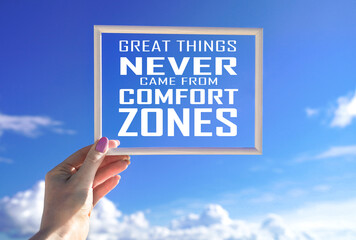 Great things never came from comfort zones. White frame in female hand with motivational words against blue sky