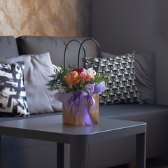 A bouquet of flowers in a paper bag with a purple ribbon stands on a gray table in a room near a...