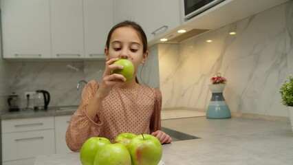 Moving shot of bunch of green apples while adorable preteen girl approaching kitchen counter, taking fruit, biting off apple, smiling, leaning on table indoors. Hungry kid having healthy snack at home