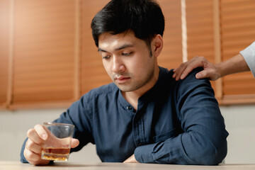 Alcoholism, depressed asian young man sitting on table while drinking alcoholic beverage, holding...