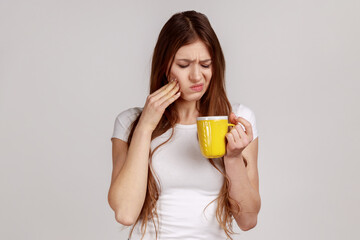 Sick unhealthy woman touching chin suffering tooth ache, has sensitive teeth, feels pain after drinking cold water, wearing white T-shirt. Indoor studio shot isolated on gray background.