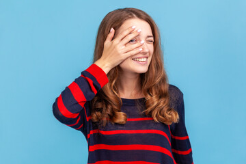 Fototapeta premium Portrait of curious young adult woman wearing sweater, peeking through fingers, looking with embarrassed inquisitive expression, watching secrets. Indoor studio shot isolated on blue background.