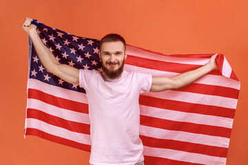 Man holding USA flag in hands, looking at camera with toothy smile, proud of his social security in country, feeling in safe, wearing pink T-shirt. Indoor studio shot isolated on orange background.