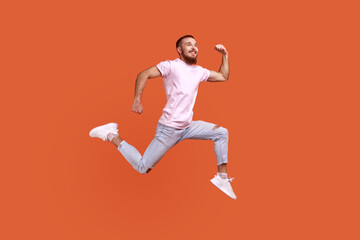 Fototapeta na wymiar Full length portrait of positive inspired bearded man jumping in air or running quickly fast, expressing happiness, wearing pink T-shirt. Indoor studio shot isolated on orange background.