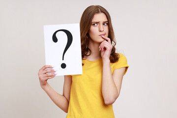 Young pensive woman in casual style attire holding paper with question mark over, thoughtful, face...