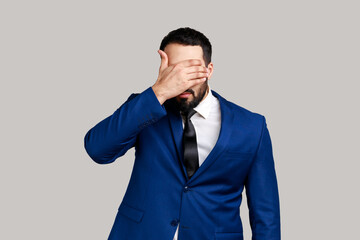 Bearded man closing eyes with hand, dont want to see that, ignoring problems, hiding from stressful situations, wearing official style suit. Indoor studio shot isolated on gray background.