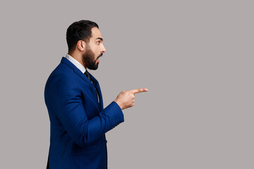Side view of bearded man looking at camera with surprised amazed expression, pointing away at copy space, wearing official style suit. Indoor studio shot isolated on gray background.