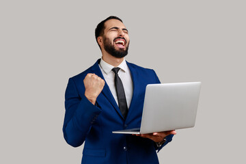 Portrait of excited bearded businessman screaming with joy and holding laptop, rejoicing victory, online betting, wearing official style suit. Indoor studio shot isolated on gray background.