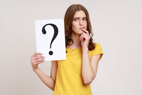 Young pensive woman in casual style attire holding paper with question mark over, thoughtful, face thinking about question, very confused idea. Indoor studio shot isolated on gray background.
