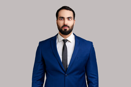Portrait of young adult bearded man looking cross-eyed, having fun with silly face expression, playing fool, wearing official style suit. Indoor studio shot isolated on gray background.