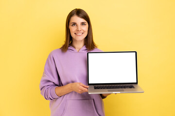 Cheerful woman standing showing laptop with white empty screen for promotion, looking at camera with smile, wearing purple hoodie. Indoor studio shot isolated on yellow background.