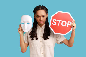 Serious woman with black dreadlocks holding white mask and red stop sign, looking at camera, do not...