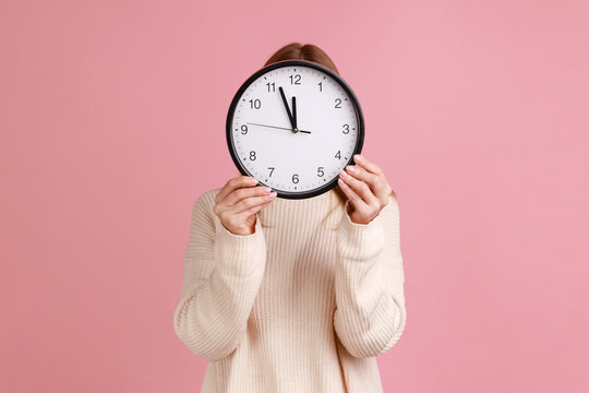 Portrait of blond woman holding wall clock hiding her face, time management, schedule and meeting appointment, wearing white sweater. Indoor studio shot isolated on pink background.