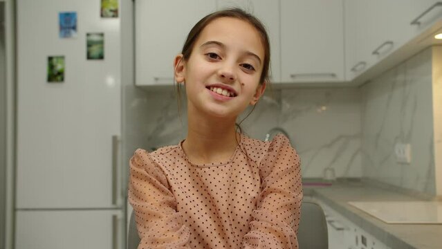 Portrait of lovely smiling girl looking directly at camera, propping chin with hands against blurred background of domestic room. Cheerful female kid expressing positive emotions indoors