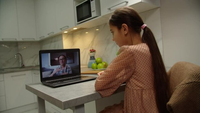 Charming school girl listening to teacher via online telecommunication app on laptop, nodding, writing down educational material to copybook at home. Moving shot. Distance classes, e-learning concept