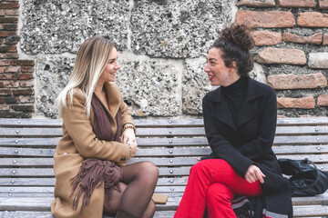 two young women sitting on a bench and chatting, young fashionable woman living the city, leisure and relax moment, soft matte filter, different female with blonde hair, brunette curly and freckles