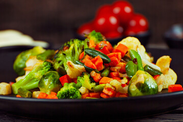 Mix of vegetables close up. Salad of various chopped vegetables. Roasted cabbage, carrots, broccoli, peas in a black plate. Food on a wooden board. Marinated tomatoes in the background.