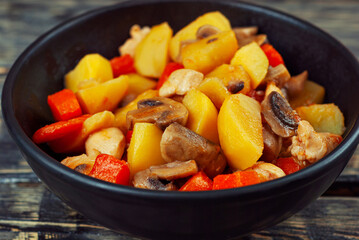Boiled potatoes with meat in a black bowl. Potatoes with mushrooms and carrots. Boiled mushrooms and chicken with potatoes. Food on a black wooden table.